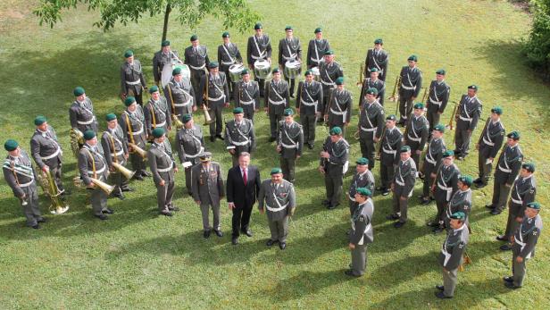 Eine Militärkapelle steht auf einer Wiese, flankiert von zwei zivil gekleideten Männern.