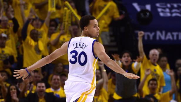 Stephen Curry mit ausgestreckten Armen im Trikot der Golden State Warriors vor jubelnden Fans.