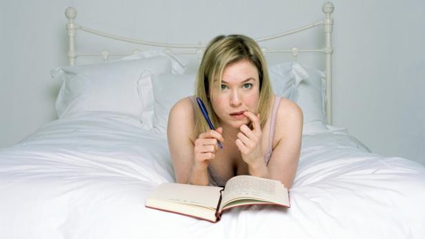 Eine Frau liegt mit einem Buch und einem Stift im Bett und scheint nachzudenken.