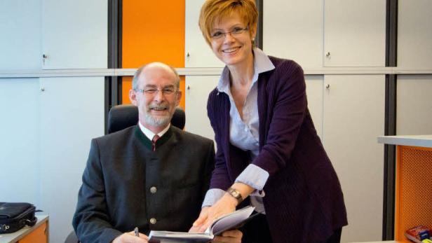 Ein Mann im Trachtenanzug sitzt am Schreibtisch, während eine Frau ihm über die Schulter schaut.