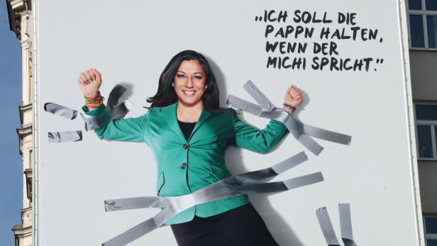 Eine Frau in einem grünen Blazer ist mit Klebeband an eine Wand geklebt, auf der ein Zitat steht.