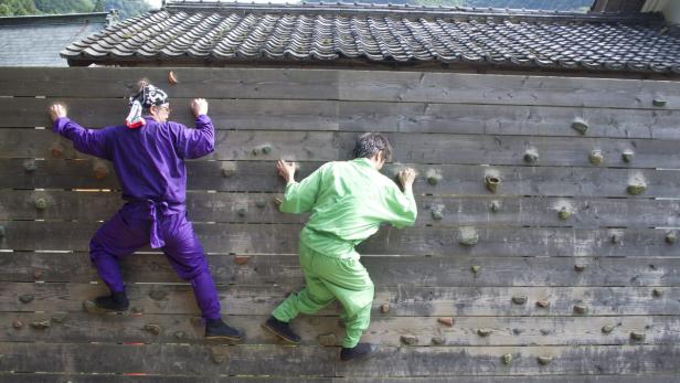 Zwei Personen in Ninja-Kostümen klettern an einer Holzwand mit Klettergriffen.