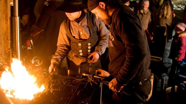 Zwei Schmiede arbeiten an einem Feuer, umgeben von Zuschauern.