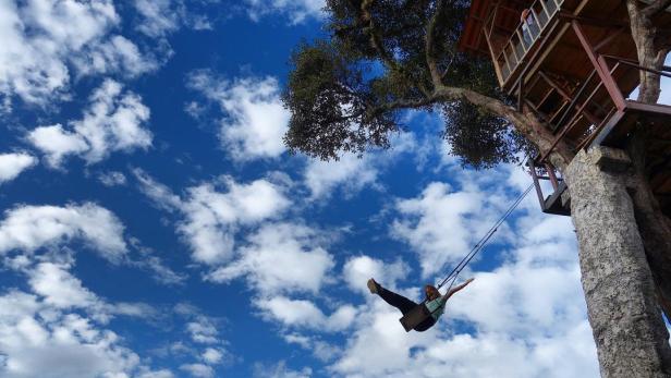 Eine Frau schwingt auf einer Schaukel vor einem Baumhaus unter blauem Himmel.