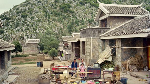 Eine Familie steht vor ihrem Haus in einem chinesischen Dorf, im Hintergrund ein bewaldeter Berg.