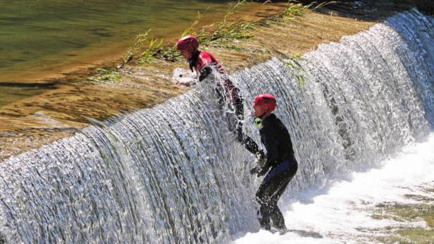 Zwei Personen in Neoprenanzügen und Helmen springen einen Wasserfall hinunter.