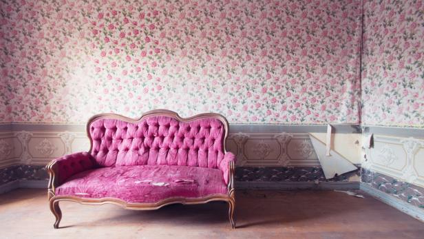 Old damaged red couch in an antique house. Flowers wallpaper in the wall