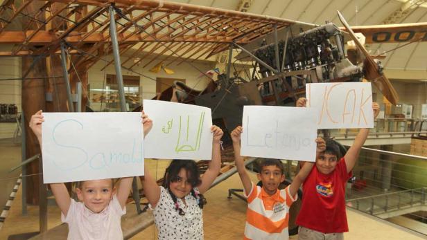 Pia, Melanie, Daniel und Ali halten Plakate mit Wörtern für Flugzeug bzw. fliegen auf Polnisch, Arabisch, Serbisch und Türkisch vor einem Flugobjekt im TMW in Kamera