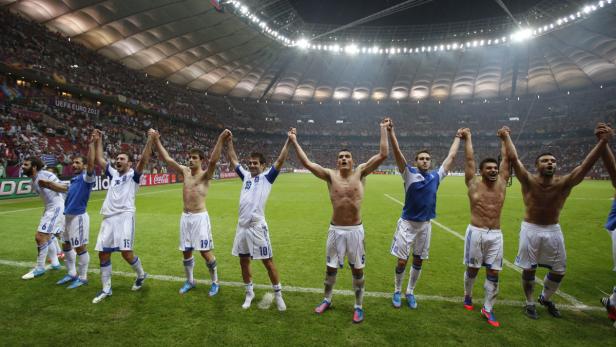 Griechische Fußballspieler feiern mit erhobenen Händen auf dem Rasen eines Stadions.
