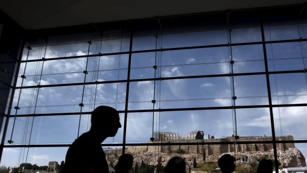 Schattenrisse von Menschen vor einer Fensterfront mit Blick auf die Akropolis in Athen.