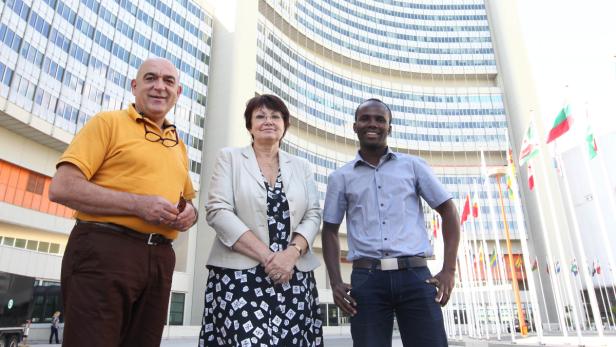 Drei Personen posieren vor dem Vienna International Centre, umgeben von Flaggen.