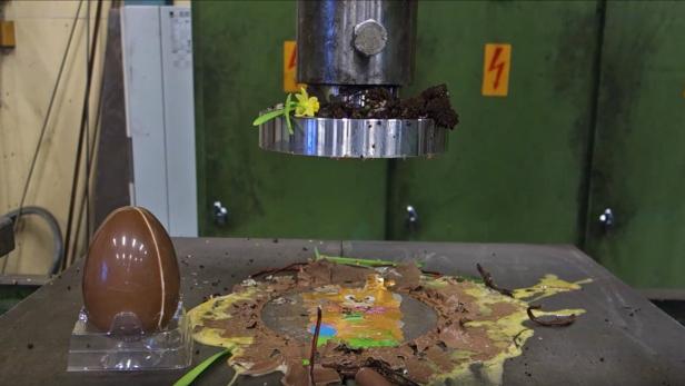 Ein Schokoladenei wird unter einer Hydraulikpresse zerdrückt.