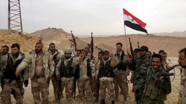 Eine Gruppe bewaffneter Soldaten posiert mit der syrischen Flagge in einer Wüstenlandschaft.
