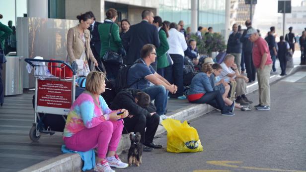 Eine Gruppe von Menschen wartet vor einem Gebäude, einige sitzen auf dem Bordstein.