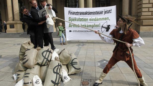 Als Robin Hood verkleideter Mann zieht an einem Seil, an dem Banker und Geldsäcke mit Dollar- und Eurozeichen hängen.