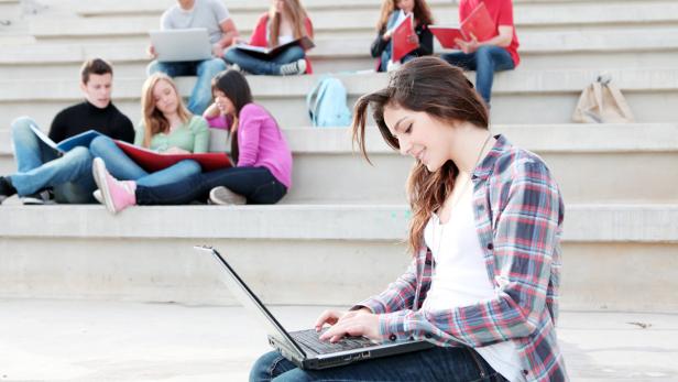 Eine junge Frau sitzt auf einer Treppe und benutzt einen Laptop, im Hintergrund weitere Studenten.