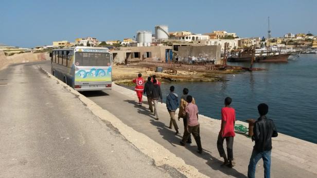 Eine Gruppe von Menschen geht an einem Hafen entlang, neben einem Bus der Organisation Misericordia.