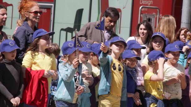 Eine Gruppe Kinder mit EU-Fahnen und blauen Kappen mit EU-Sternen bei einer Veranstaltung.