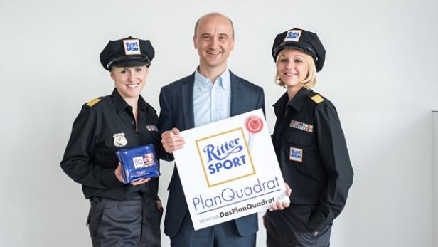 Zwei Frauen in Uniform und ein Mann präsentieren eine Tafel von Ritter Sport und ein Schild von „PlanQuadrat“.