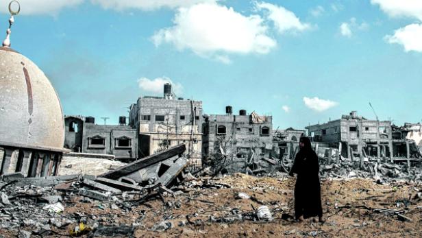 Eine Frau steht vor einer zerstörten Moschee und Trümmern in einer Stadt.
