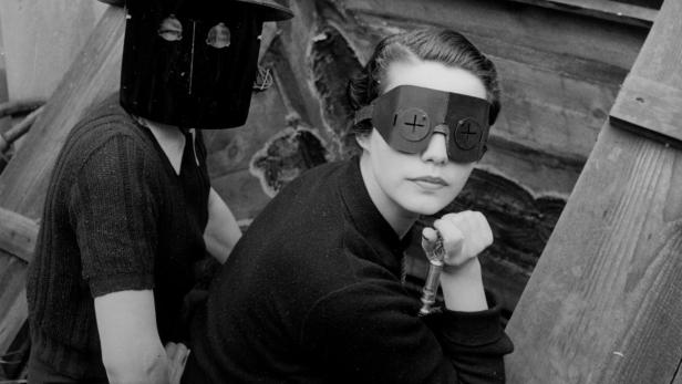 Zwei Personen in ungewöhnlichen Masken posieren vor einer Holzkulisse.