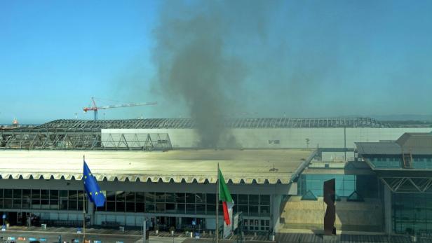Rauch steigt über dem Flughafen von Pisa auf, im Vordergrund wehen die Europa- und Italienflagge.