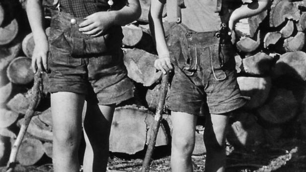 Zwei barfüßige Jungen in Lederhosen posieren vor einem Holzstapel.