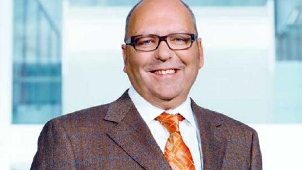 Ein lächelnder Mann mit Brille und Anzug steht vor einem hellen Hintergrund.