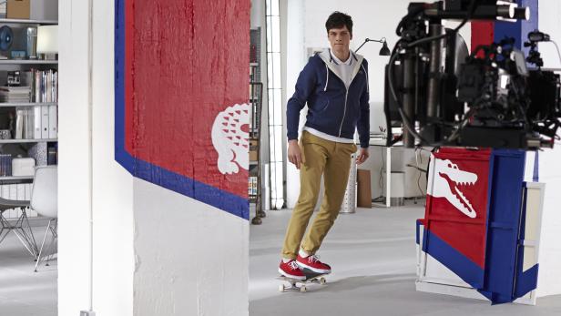 Ein Mann auf einem Skateboard in einem Studio mit Requisiten im Lacoste-Stil.