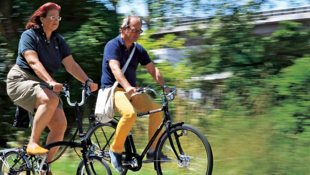 Ein älteres Paar fährt mit Fahrrädern der Marke Puch durch eine grüne Landschaft.
