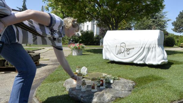 Eine Frau beugt sich über ein Grab mit Kerzen und Blumen, im Hintergrund ein verhülltes Denkmal.