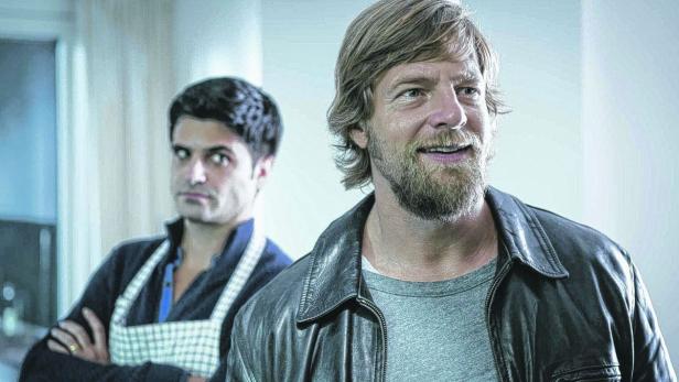 Zwei Männer stehen in einem Raum, einer mit Kochschürze, der andere mit Lederjacke.