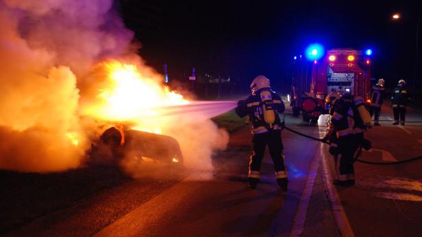 Feuerwehrleute löschen ein brennendes Auto auf einer dunklen Straße.