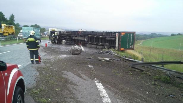 Ein umgekippter Lastwagen liegt auf der Seite, während Feuerwehrleute am Unfallort stehen.