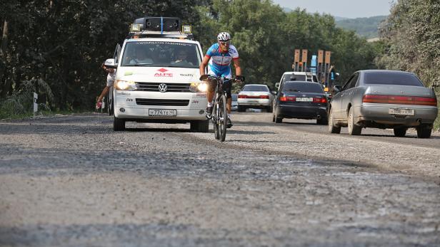 Ein Radfahrer fährt auf einer unbefestigten Straße, begleitet von mehreren Fahrzeugen.