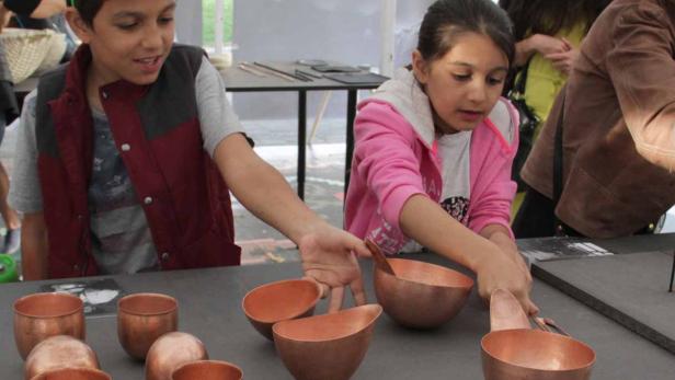 Zwei Kinder betrachten und berühren Kupferschalen auf einem Tisch.
