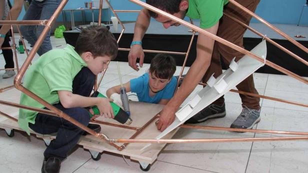 Zwei Jungen und ein Mann arbeiten an einer Konstruktion aus Kupferrohren.