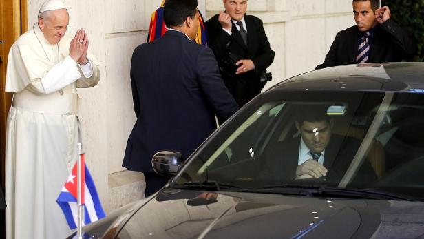 Papst Franziskus steht neben einer kubanischen Flagge und begrüßt mit gefalteten Händen.