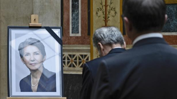 Ein Porträt einer Frau mit einer Perlenkette steht auf einer Staffelei, umgeben von Trauergästen.