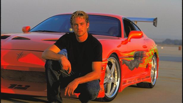 Paul Walker posiert vor einem roten Toyota Supra mit auffälligen Aufklebern.