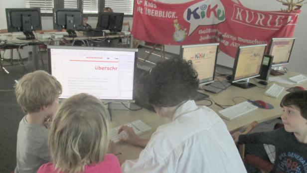 Kinder und eine Frau arbeiten an Computern in einem Raum mit einem „KiKu Kurier“-Banner.