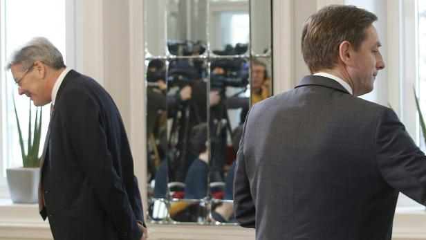 Zwei Männer in Anzügen gehen an einem Spiegel vorbei, in dem sich Kameraleute spiegeln.