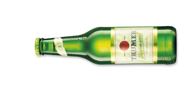 Eine grüne, geöffnete Flasche Trumer Pils liegt auf einer weißen Oberfläche.
