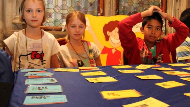 Drei Kinder sitzen an einem Tisch mit Karten und einem Cartoon im Hintergrund.