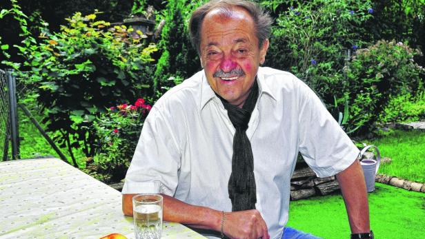 Ein lächelnder älterer Mann mit Schnurrbart sitzt an einem Tisch im Garten.