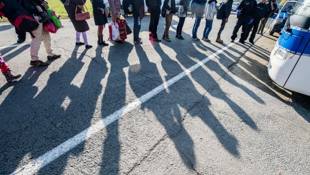 Eine Schlange von Menschen steht neben einem Polizeifahrzeug, ihre Schatten fallen auf den Asphalt.
