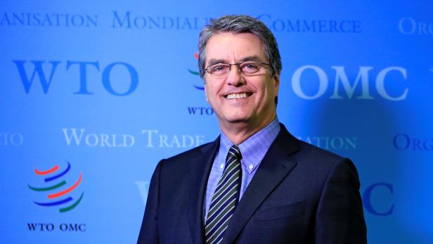 Ein Mann mit Brille und Anzug lächelt vor einem WTO-Hintergrund.