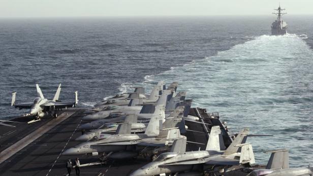 Mehrere Kampfjets stehen auf dem Deck eines Flugzeugträgers, während im Hintergrund ein Kriegsschiff fährt.