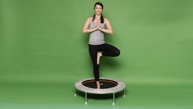 Eine Frau macht Yoga auf einem Minitrampolin vor einem grünen Hintergrund.