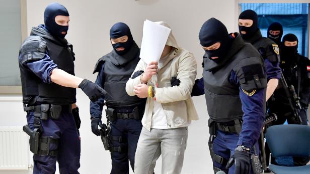 Polizisten in Kampfmontur führen einen Mann mit Handschellen ab, der sein Gesicht mit einem Blatt Papier bedeckt.